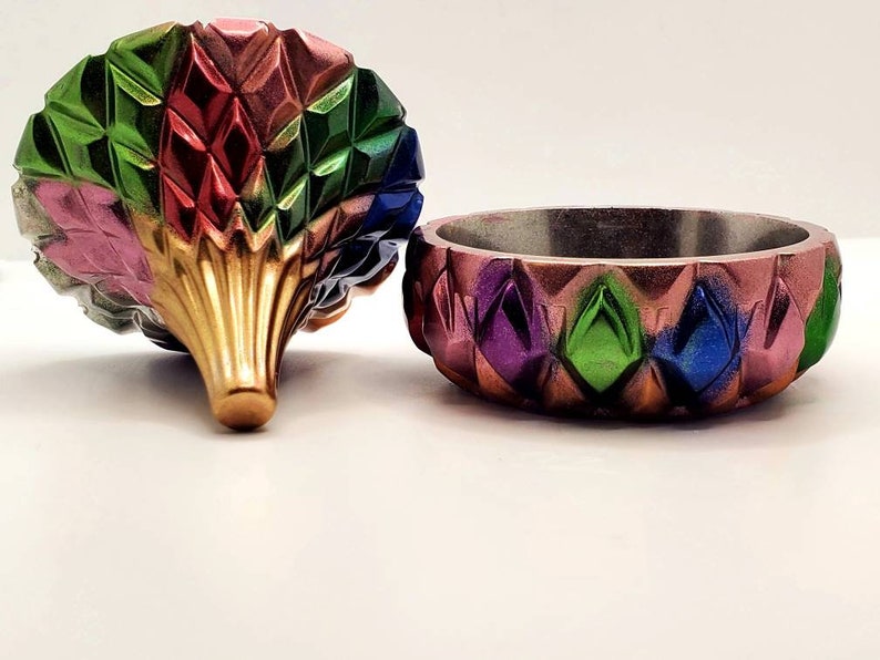 May include: A colorful, geometric trinket box with a lid shaped like a flower. The box is made of resin and has a shiny, metallic finish. The lid is decorated with a pattern of diamonds in shades of pink, red, green, blue, and gold.