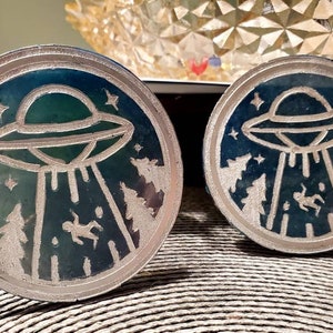 May include: Two round, silver-colored coasters with a blue and green design. The design features a white silhouette of a UFO with beams of light shining down on a forest and a figure falling from the sky.