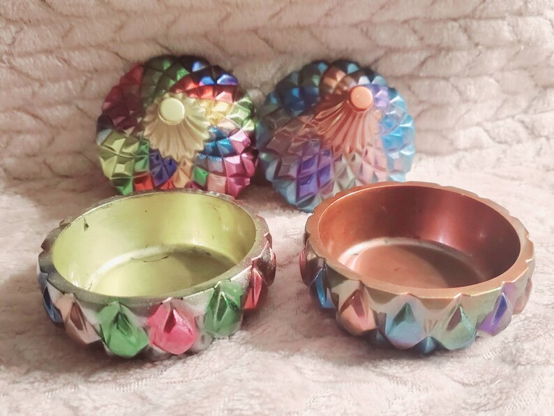 May include: Two small, round, iridescent trinket boxes with lids. The lids are decorated with a geometric pattern of colorful, iridescent triangles. The boxes are open and the inside of each box is a different color. One box is green and the other is brown.