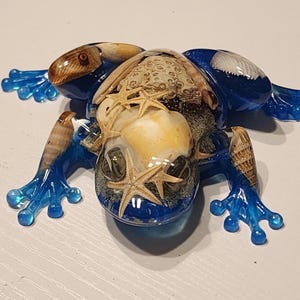 May include: A blue glass frog figurine with seashells and starfish embedded inside. The frog is sitting on a white surface.