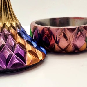 May include: A colorful, geometric bowl and lid with a diamond pattern. The bowl is made of metal and has a pink, orange, red, and green color scheme. The lid is made of metal and has a purple, gold, and blue color scheme.