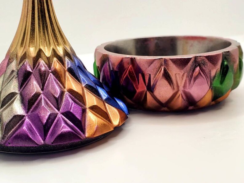 May include: A colorful, geometric bowl and lid with a diamond pattern. The bowl is made of metal and has a pink, orange, red, and green color scheme. The lid is made of metal and has a purple, gold, and blue color scheme.
