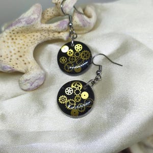 Peut inclure: Une paire de boucles d'oreilles de style steampunk. Chaque boucle d'oreille présente un pendentif circulaire noir avec des engrenages et des éléments mécaniques dorés. Les boucles d'oreilles sont suspendues à des crochets d'oreille argentés. Le fond est un tissu blanc.