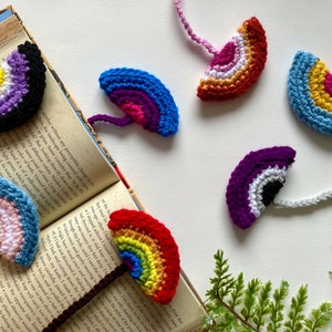 Puede incluir: Seis marcapáginas de ganchillo con forma de arcoíris en diferentes colores, incluyendo morado, azul, rosa, naranja, amarillo y rojo. Los marcapáginas están colgados de un libro.