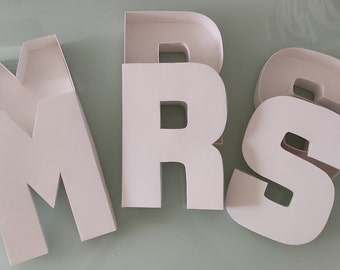 May include: White cardboard letters spelling out the word "MRS" in a decorative font. The letters are hollow and have a three-dimensional appearance.