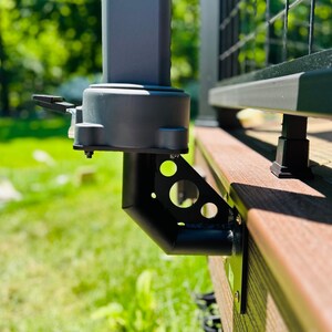 May include: A matte black metal mounting bracket attached to the side of a wooden deck, featuring a triangular support plate with circular cutouts. The bracket supports a dark gray vertical post base with a small black lever on the side.