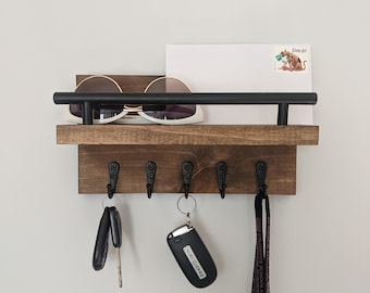 Può includere: Un porta chiavi in legno con un ripiano in metallo nero e cinque ganci. Sul ripiano ci sono un paio di occhiali da sole e una busta bianca con un adesivo di Scooby Doo. Ci sono due set di chiavi e un laccio che pendono dai ganci.