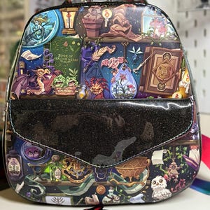 May include: A black glittery backpack with a colorful fantasy illustration. The design features various magical creatures, books, and potions. The flap has a black glittery finish, and the bag includes a top handle and adjustable straps.