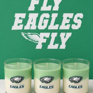 May include: Three glass candles with layered light green and off-white wax. Each candle features an Eagles logo and the word EAGLES printed on the front. The background displays the white text FLY EAGLES FLY on a solid green surface.