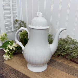 May include: A white ceramic coffee pot with a lid and a curved handle. The pot has a rounded base and a long spout. The pot is decorated with vertical lines and sits on a wooden surface.