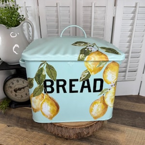May include: A light blue bread box with a lid and handle, decorated with watercolor lemons and green leaves. The word "BREAD" is printed in large black letters on the front. The bread box is sitting on a wooden surface.