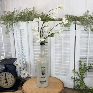 May include: A light beige ceramic bottle vase featuring a black rectangular label with botanical illustrations and text. The bottle has a narrow neck and a black rim, and it contains a branch of white magnolia flowers with green leaves.