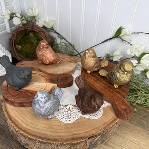 May include: A collection of decorative bird figurines in various metallic finishes, including copper, gold, silver, and bronze. The birds are displayed on wooden platforms and a decorative cutting board, with a rustic and natural aesthetic.