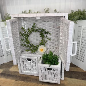 May include: A white wicker storage chest with two small drawers and a square planter. The chest is open, revealing a green wreath and a white flower. The chest has a handle on each side.
