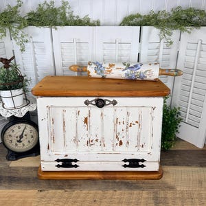 May include: A rectangular wooden bread box with a distressed white finish and a natural wood top. A rolling pin with a blue bird and floral pattern rests on top. The bread box features a black metal handle and two black hinges on the front.
