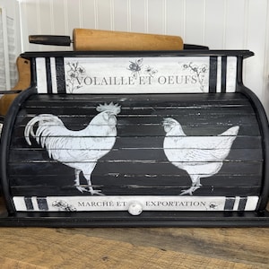 May include: Black and white bread box with a rooster and hen design. The box has the words "VOLAILLE ET OEUFS" and "MARCHÉ ET EXPORTATION" printed on it. A wooden rolling pin rests on top of the box.