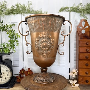 May include: A copper-colored metal urn with a hammered texture on the upper rim, a central embossed floral medallion, and two curved side handles. The vessel features a pedestal base with leaf-like detailing and a smooth, polished finish.