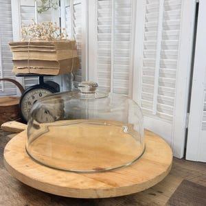 May include: A wooden serving board with a clear glass dome. The board is round with a natural wood finish. The glass dome has a small knob on top. The background includes a vintage scale and a small basket.