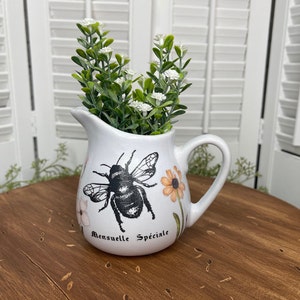 May include: White ceramic pitcher with a black bee and floral design. The pitcher has the text "Mensuelle Speciale" on the bottom. The pitcher is filled with white flowers.