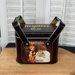 May include: A black metal tin with a handle, featuring a vintage illustration of a child holding a cup of Hershey's Cocoa. The tin has the text "Hershey's Cocoa" on the front and "Hershey's Chocolate" on the bottom.