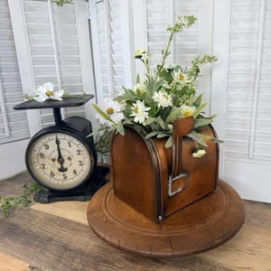 May include: A copper-colored metal mailbox container filled with a bouquet of white and green artificial flowers, positioned on a round wooden base next to a black vintage-style scale with a white face and a single white flower on top.