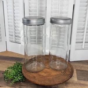 May include: Two clear glass jars with silver lids sit on a round, wooden tray. The jars are tall and cylindrical with a textured surface. The background features white, slatted shutters and a small green plant.