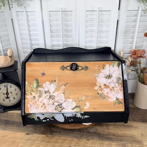 May include: A black wooden bread box with a natural wood front panel featuring a floral print of light pink flowers and a small bee. The box has a black metal handle and a hinged door that opens to reveal the interior storage space.
