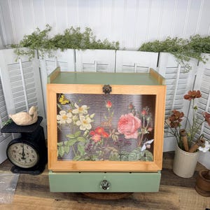May include: A sage green wooden bread box with a light wood frame and a ribbed glass front panel featuring a colorful floral print of roses and white flowers. The box includes a single bottom drawer with a clear glass knob and a metal latch.