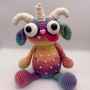 Puede incluir: Un animal de peluche de crochet arcoíris con un cuerno de unicornio, ojos saltones y una lengua rosa que sobresale. El animal está sentado sobre una superficie blanca.