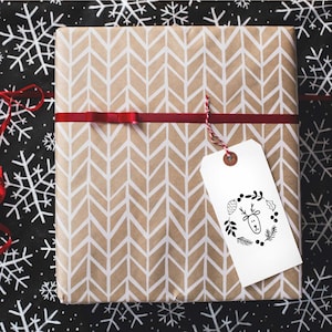 Pu&ograve; includere: Una scatola regalo marrone con un motivo a chevron e un'etichetta regalo bianca con un'illustrazione in bianco e nero di una renna con una ghirlanda intorno alla testa. L'etichetta regalo ha il testo "Buon Natale".