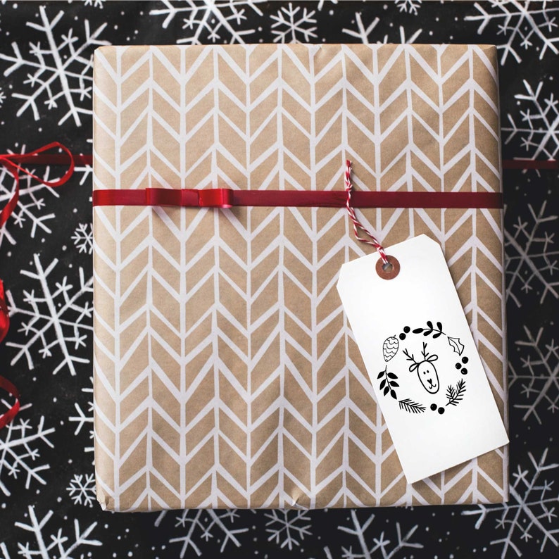 Pu&ograve; includere: Una scatola regalo marrone con un motivo a chevron e un'etichetta regalo bianca con un'illustrazione in bianco e nero di una renna con una ghirlanda intorno alla testa. L'etichetta regalo ha il testo "Buon Natale".