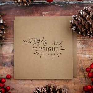 Pu&ograve; includere: Un biglietto di carta kraft marrone con il testo "merry & BRIGHT" in font calligrafico nero. Il biglietto &egrave; circondato da pigne e bacche rosse.
