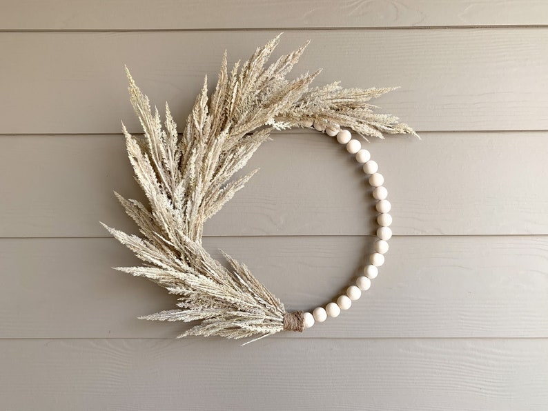 May include: A wreath made of dried pampas grass and white wooden beads. The wreath is a natural color and is hanging on a light brown wall.