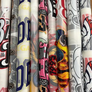 Pu&ograve; includere: Una collezione di otto rotoli di adesivi in vinile con vari design, tra cui un'auto rosa, un cane dei cartoni animati con occhiali da sole, un logo rosso e bianco con la parola "RAIDERS" e un cavallo bianco e nero.