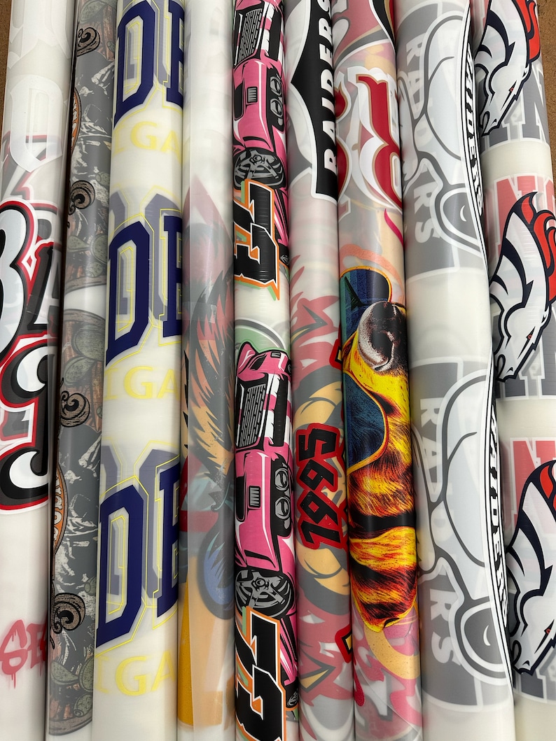 Pu&ograve; includere: Una collezione di otto rotoli di adesivi in vinile con vari design, tra cui un'auto rosa, un cane dei cartoni animati con occhiali da sole, un logo rosso e bianco con la parola "RAIDERS" e un cavallo bianco e nero.