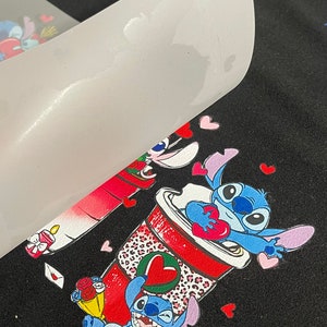 Pu&ograve; includere: Una maglietta nera con un design colorato con due personaggi dei cartoni animati blu che tengono cuori. I personaggi sono circondati da cuori rosa. Il design &egrave; stampato su un foglio di trasferimento bianco.