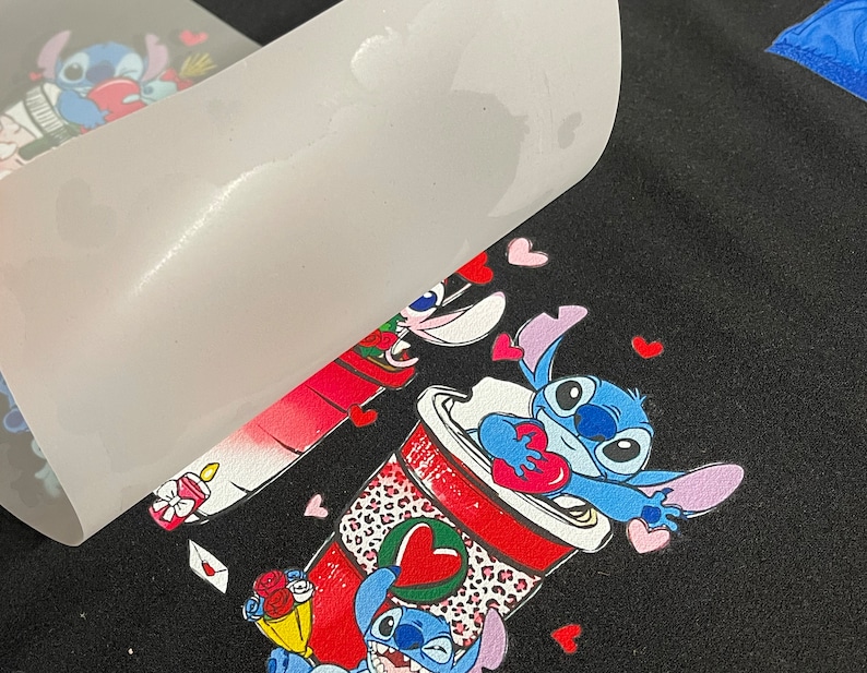 Pu&ograve; includere: Una maglietta nera con un design colorato con due personaggi dei cartoni animati blu che tengono cuori. I personaggi sono circondati da cuori rosa. Il design &egrave; stampato su un foglio di trasferimento bianco.