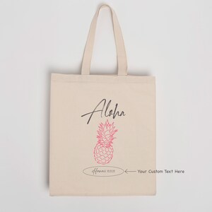 May include: A beige tote bag with a pink pineapple design and the word "Aloha" in black script. The bag has a white oval with the text "Hawaii 01.22.22" and an arrow pointing to the right with the text "Your Custom Text Here".