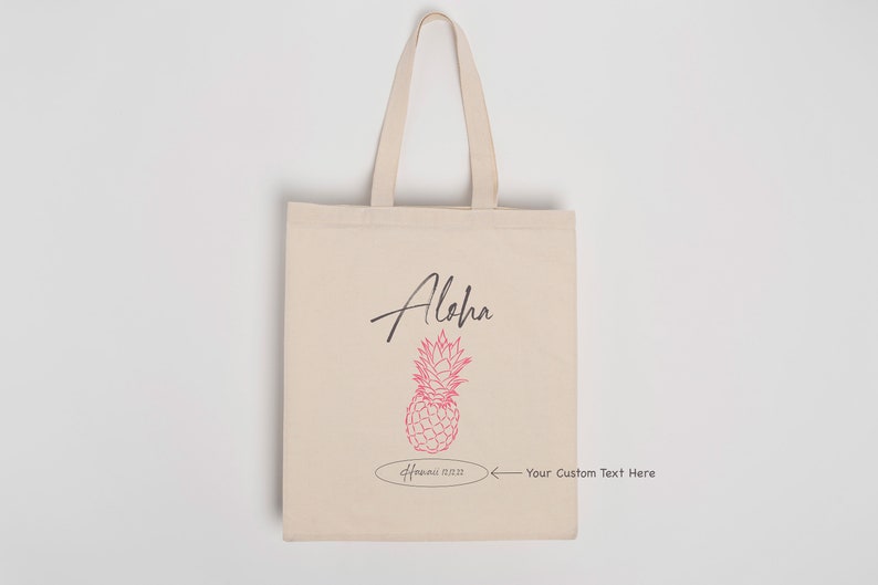 May include: A beige tote bag with a pink pineapple design and the word "Aloha" in black script. The bag has a white oval with the text "Hawaii 01.22.22" and an arrow pointing to the right with the text "Your Custom Text Here".