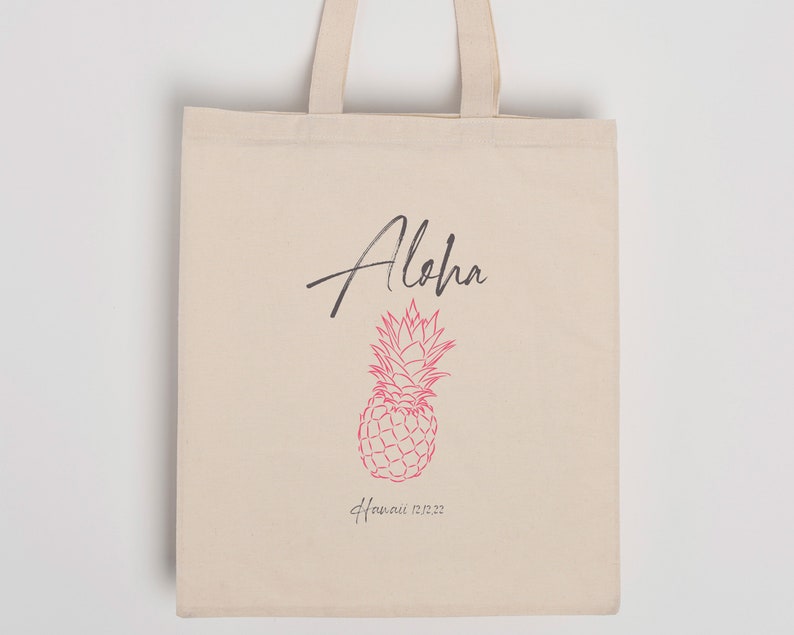 May include: A white canvas tote bag with black handles. The bag has a pink pineapple design and the text "Aloha" in black script and "Hawaii 12.22" in black text.