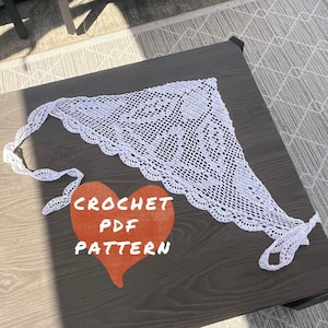 May include: A white, crocheted triangle scarf with a scalloped edge, lying on a dark wooden surface. The scarf has a delicate, openwork design. A red heart shape with the words "CROCHET PDF PATTERN" is below the scarf.