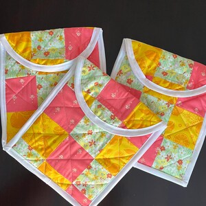 May include: Three quilted fabric pouches with a patchwork design. The pouches feature a mix of pink, yellow, and green floral and geometric patterns. Each pouch has a white trim and a curved flap.