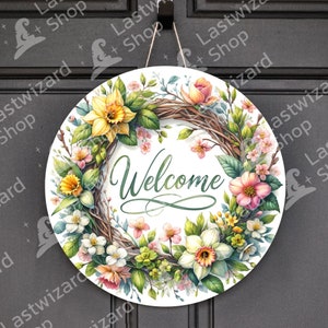 Op de afbeelding: Een ronde welkomstkrans met een witte achtergrond. De krans is gemaakt van gevlochten takken en is versierd met verschillende bloemen, waaronder narcissen, roze bloemen en witte bloemen. Het woord "Welkom" staat in groene cursieve letters in het midden van de krans.
