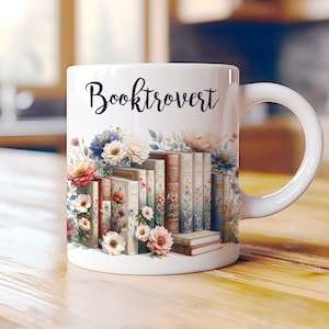 Puede incluir: Taza de cerámica blanca con un diseño floral y una pila de libros. La taza tiene el texto "Booktrovert" impreso en ella.