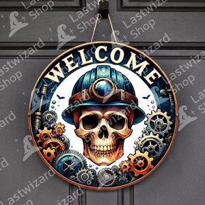 May include: Round wooden welcome sign featuring a skull wearing a miner's helmet, surrounded by gears. The word "WELCOME" is displayed in white. The sign is predominantly brown, blue, and orange, with a hanging string.