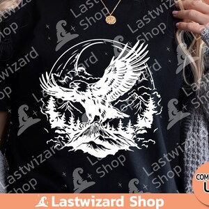 May include: A black t-shirt with a white graphic of an eagle soaring over a mountain range. The eagle is in flight with its wings spread wide. The mountain range is in the background and is made up of several peaks. The graphic is surrounded by a circle of clouds.