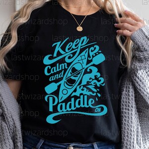 May include: Black t-shirt with a blue graphic design that says "Keep Calm and Paddle On". The graphic features a kayak with paddles and waves.