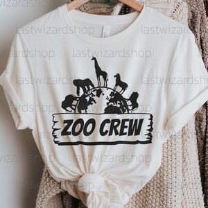 Pu&ograve; includere: Una maglietta bianca con una grafica nera di un globo circondato da animali. Il testo "ZOO CREW" &egrave; su un cartello di legno sotto il globo.