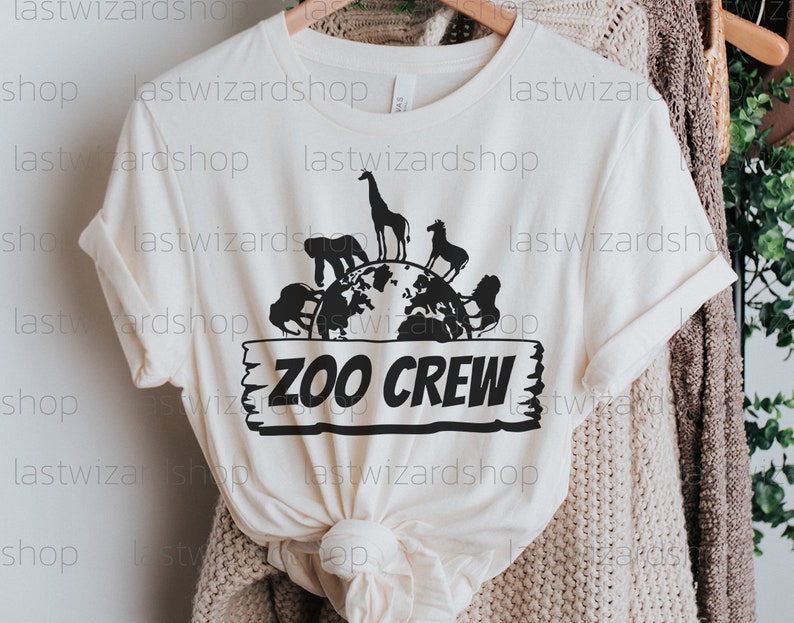 Pu&ograve; includere: Una maglietta bianca con una grafica nera di un globo circondato da animali. Il testo "ZOO CREW" &egrave; su un cartello di legno sotto il globo.