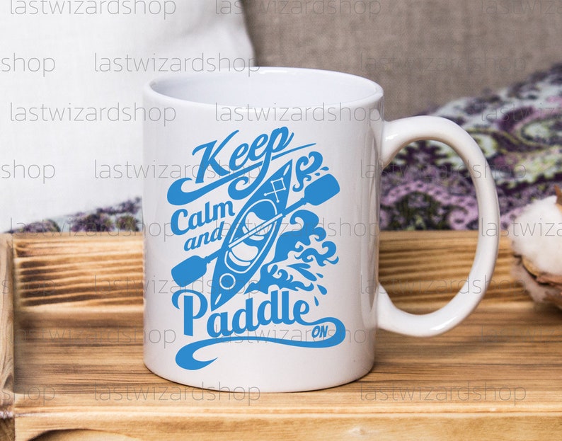 May include: White ceramic mug with a blue graphic that says "Keep Calm and Paddle On" with a stylized image of a kayak and paddle.